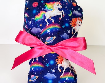 May include: A blue scrub hat with a pink ribbon tied around it. The hat has a pattern of unicorns, rainbows, stars, and planets on a dark blue background.
