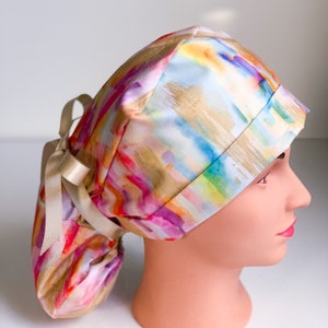 May include: A colorful surgical scrub hat with a watercolor-style print in shades of pink, orange, yellow, green, blue, and purple. The hat has a long ponytail opening and a gold ribbon tie.