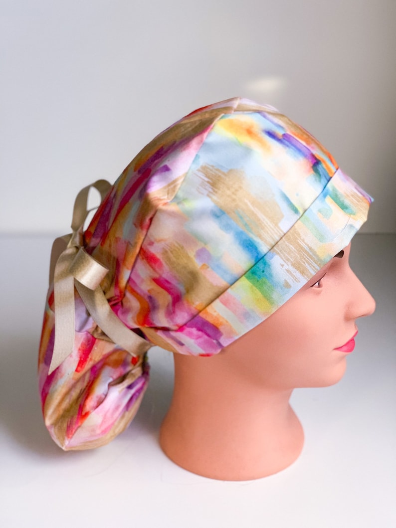 May include: A colorful surgical scrub hat with a watercolor-style print in shades of pink, orange, yellow, green, blue, and purple. The hat has a long ponytail opening and a gold ribbon tie.