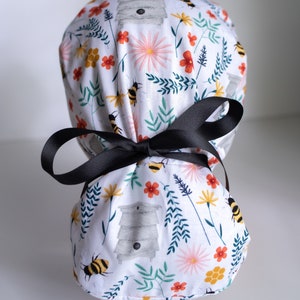 May include: White scrub hat with a floral pattern featuring bees, flowers, and beehives. The hat has a black ribbon tie.