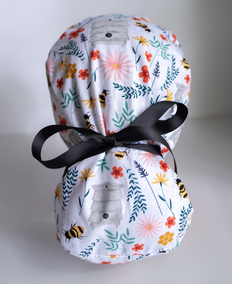 May include: White scrub hat with a floral pattern featuring bees, flowers, and beehives. The hat has a black ribbon tie.