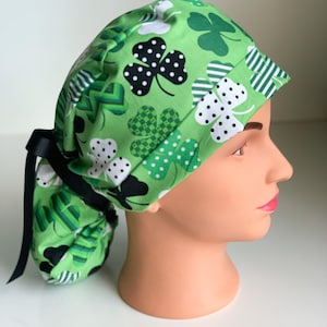 May include: Green scrub cap with a St. Patrick's Day theme. The cap features a pattern of shamrocks in various shades of green, some with white polka dots. A black ribbon is tied at the back. The cap is designed to be worn by medical professionals.
