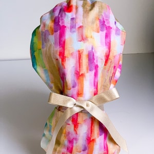 May include: A colorful surgical scrub hat with a watercolor-like pattern of pink, orange, yellow, and purple brushstrokes. The hat is tied with a gold satin ribbon.