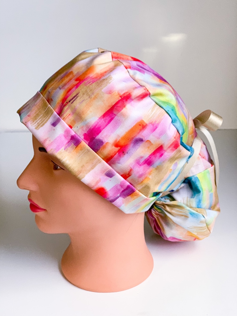 May include: A colorful surgical scrub hat with a watercolor-like pattern of pink, orange, yellow, and blue stripes. The hat has a tie at the back.