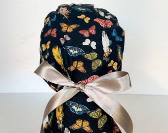 Könnte beinhalten: Eine schwarze OP-Haube mit einem Muster aus bunten Schmetterlingen, Federn und floralen Elementen. Sie ist mit einem beigen Satinband gebunden. Das Design umfasst Schmetterlinge in Gelb-, Orange-, Blau- und Rosatönen.