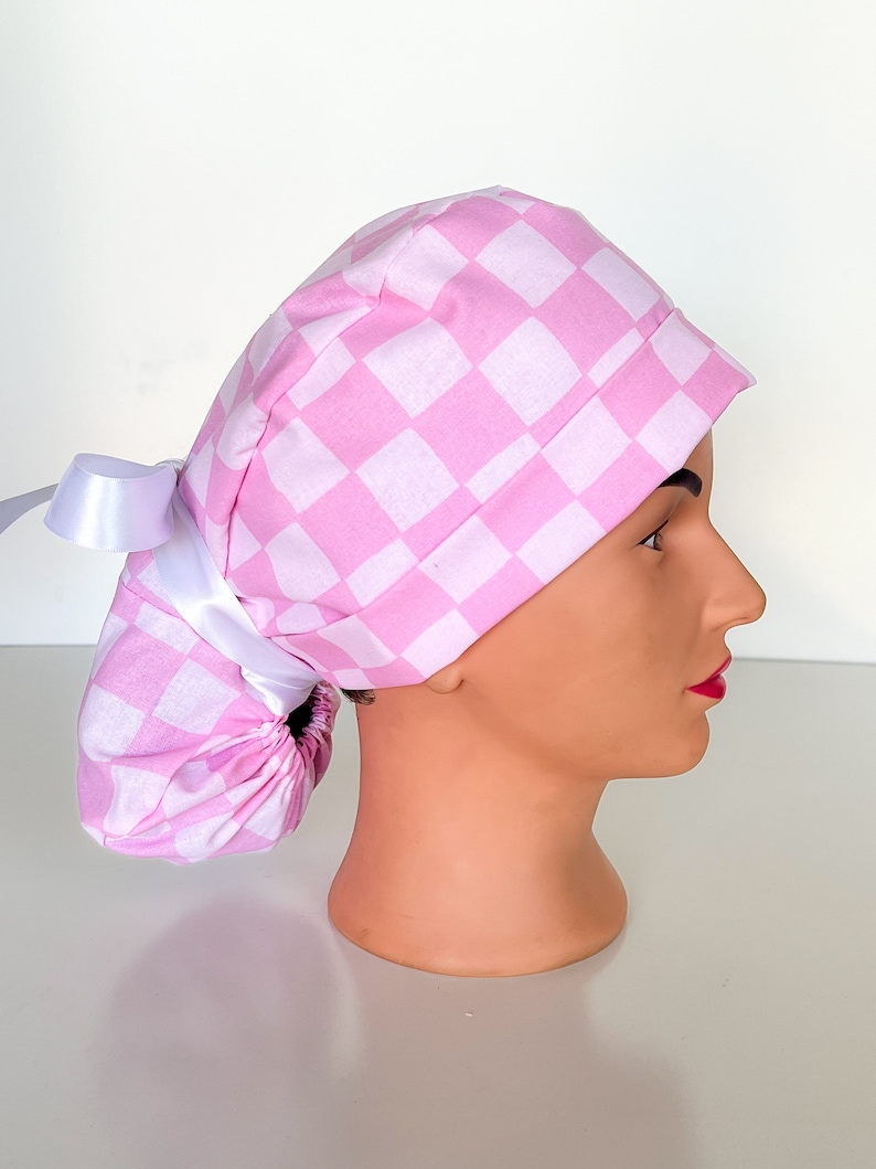 May include: Pink and white checkered surgical scrub hat with a white satin tie. The hat is designed to be worn with a ponytail.