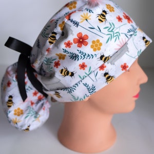 May include: A white scrub hat with a floral and bee print. The hat has a black ribbon tie and a gathered back.