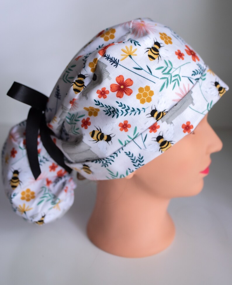 May include: A white scrub hat with a floral and bee print. The hat has a black ribbon tie and a gathered back.