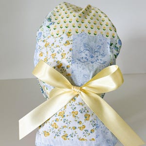 May include: A scrub cap featuring a patchwork design of floral and damask patterns in shades of yellow, white, and light blue. The fabric is tied at the back with a smooth, pale yellow satin ribbon, creating a gathered and rounded silhouette.