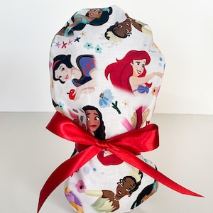 May include: A white fabric scrub hat with a red ribbon tied around it. The hat features a pattern of Disney princesses, including Snow White, Ariel, Tiana, Jasmine, and Pocahontas.