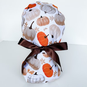 May include: A white scrub cap featuring a repeating pattern of pumpkins in shades of orange, brown, and gray, with autumn leaves. The cap is tied with a dark brown satin bow.