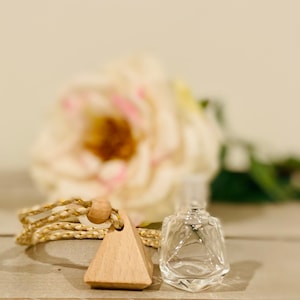 May include: A wooden triangle diffuser with a gold and white cord and a clear glass bottle with a gold lid. The diffuser is hanging from the cord and the bottle is sitting on a wooden surface. A white rose is in the background.