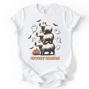 May include: White t-shirt featuring a stack of three capybaras with a pumpkin, ghosts, and bats. The text "Spooky Season" is printed in orange below the capybaras. A fun, Halloween-themed graphic tee.