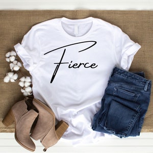 May include: White t-shirt with the word "Fierce" written in black cursive script. The shirt is laid flat on a brown hessian surface with a pair of brown ankle boots and blue jeans.