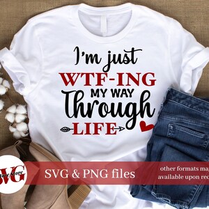 May include: White t-shirt with black text that reads "I'm just WTF-ING my way through LIFE" with a red heart and arrow. The shirt is on a brown surface with a pair of jeans and a cotton ball.