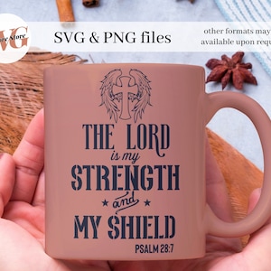 以下が含まれることがあります： 翼のある十字架と「THE LORD is my STRENGTH and MY SHIELD PSALM 28:7」というテキストが描かれた青いデザインのピンクのセラミックマグカップです。