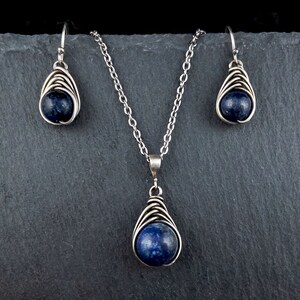 Pode incluir: Um conjunto de colar e brincos de prata com um pingente de pedra preciosa azul escuro e brincos combinando. O pingente e os brincos s&atilde;o envoltos em arame e t&ecirc;m a forma de uma l&aacute;grima.