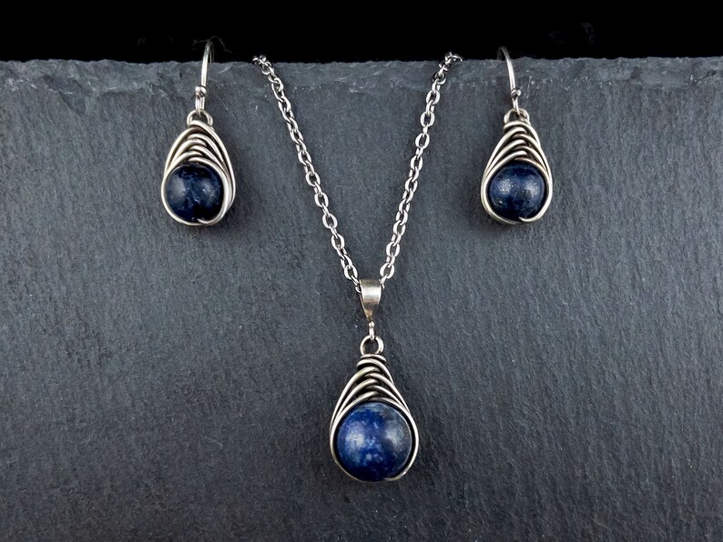 Pode incluir: Um conjunto de colar e brincos de prata com um pingente de pedra preciosa azul escuro e brincos combinando. O pingente e os brincos s&atilde;o envoltos em arame e t&ecirc;m a forma de uma l&aacute;grima.