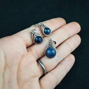 Pode incluir: Um conjunto de brincos e um pingente de prata com uma pedra preciosa azul escura envolvida em fio de prata.