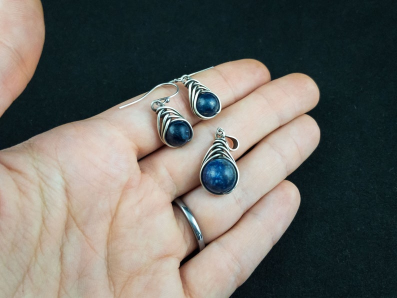 Pode incluir: Um conjunto de brincos e um pingente de prata com uma pedra preciosa azul escura envolvida em fio de prata.