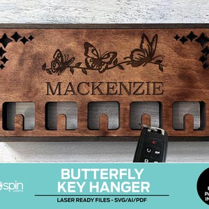 Puede incluir: Un colgador de llaves de madera con acabado marrón oscuro, con diseños de mariposas cortados con láser y el nombre "MACKENZIE". Tiene ranuras para llaves y recortes decorativos. Una llave de coche está insertada en una de las ranuras. El texto "BUTTERFLY KEY HANGER" está en la parte inferior.