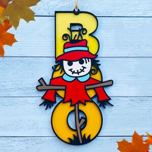 May include: A yellow wooden letter "B" with a scarecrow face and red hat. The scarecrow is holding a stick with a brown and yellow striped pattern. The letter is hanging on a light blue wooden background.