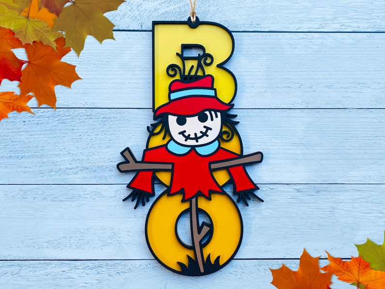 May include: A yellow wooden letter "B" with a scarecrow face and red hat. The scarecrow is holding a stick with a brown and yellow striped pattern. The letter is hanging on a light blue wooden background.