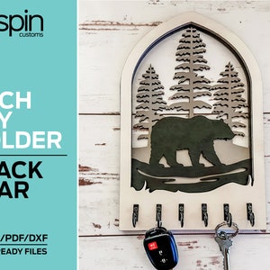 May include: A wooden key holder shaped like an arched doorway with a black bear and pine trees inside. The key holder has six hooks for hanging keys. The text "ARCH KEY HOLDER BLACK BEAR" is printed on the left side of the image. The text "SVG/AI/PDF/DXF LASER READY FILES" is printed below the key holder.
