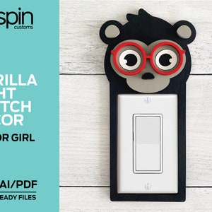 May include: A wooden light switch cover with a gorilla face design. The gorilla has black fur, red glasses, and a gray nose. The light switch cover is laser cut and ready to be assembled.