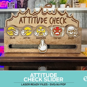 以下が含まれることがあります： さまざまな絵文字の表現と「ATTITUDE CHECK」の文字が入った木製の「Attitude Check」スライダー。 スライダーには中指のアイコンと「YA,OK」、「JUST NO」、「WHATEVER」、「TRY ME」、「FAAF」のフレーズがあります。