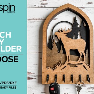 May include: Wooden key holder shaped like an arch with a moose silhouette inside. The key holder has five hooks and is laser cut. The text "spin customs" is in the top left corner. The text "ARCH KEY HOLDER MOOSE" is in the top left corner. The text "SVG/AI/PDF/DXF LASER READY FILES" is in the bottom left corner.