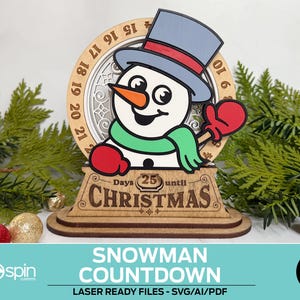 May include: A wooden snowman countdown decoration with a light blue hat, red gloves, and a green scarf. The countdown shows "25 days until Christmas". The design includes a circular frame with numbers and the text "Snowman Countdown".
