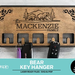 May include: Wooden key hanger with a bear family design, personalized with "The Mackenzie Family." The key holder has four key slots and decorative corner accents. The words "BEAR KEY HANGER" are at the bottom.