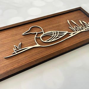 May include: A wooden wall art piece featuring a stylized loon with its wings spread, resting on a branch with reeds. The loon and reeds are cut out of a lighter wood and mounted on a dark brown wood background.