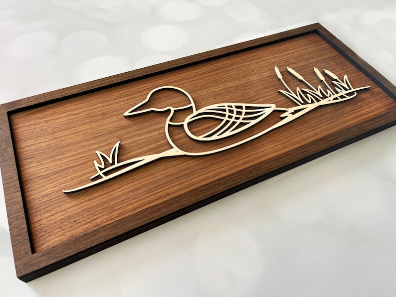 May include: A wooden wall art piece featuring a stylized loon with its wings spread, resting on a branch with reeds. The loon and reeds are cut out of a lighter wood and mounted on a dark brown wood background.