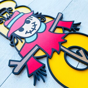 May include: A yellow number three with a black outline. A scarecrow with a red shirt, blue collar, and brown hat is standing in front of the number. The scarecrow has a stitched smile and black eyes.