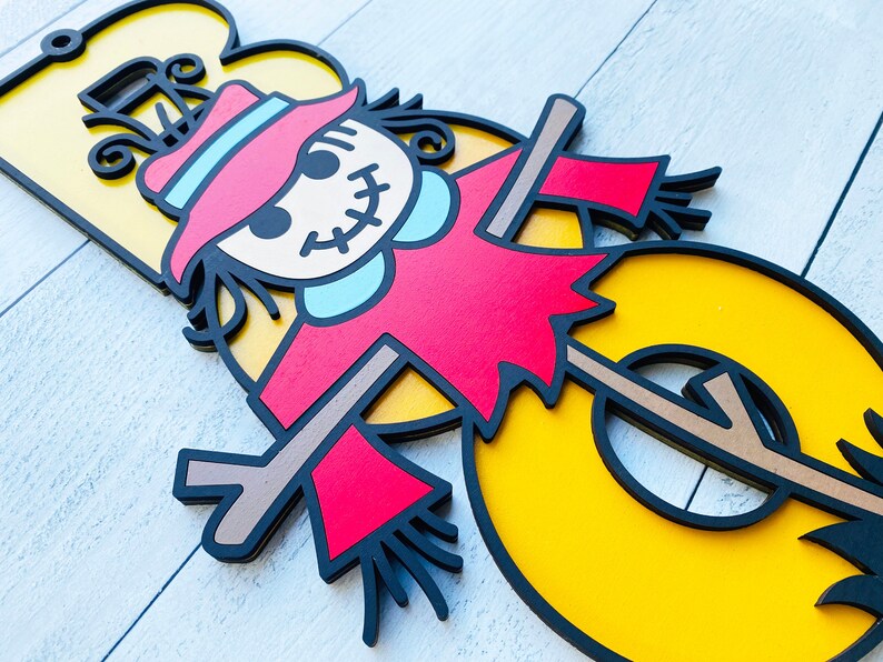 May include: A yellow number three with a black outline. A scarecrow with a red shirt, blue collar, and brown hat is standing in front of the number. The scarecrow has a stitched smile and black eyes.