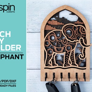 Op de afbeelding: Houten sleutelhanger in de vorm van een boog met een olifantontwerp. De olifant is ingewikkeld gesneden met een bloemmotief en heeft een donkerbruine vlek. De sleutelhanger heeft vijf haken om sleutels aan te hangen.