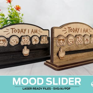 Può includere: Due cursori di umore in legno con la scritta "TODAY I AM" sopra una fila di faccine emoji. Un cursore a forma di mano indica l'umore attuale. Uno è marrone scuro, l'altro è marrone chiaro. Il testo "MOOD SLIDER" è in basso.
