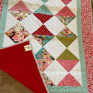 Puede incluir: Un edredón hecho a mano con un diseño de patchwork. El edredón presenta cuadrados blancos y formas de diamante coloridas en rojo, rosa, verde y estampados florales. El borde es rojo con un diseño floral blanco, y el reverso es rojo liso.