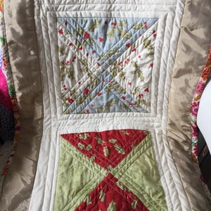 Puede incluir: Primer plano de una funda de silla acolchada con un diseño de patchwork. La funda presenta cuadrados de tela con varios patrones, incluyendo hojas y bayas de acebo sobre un fondo azul claro y rojo. Los bordes son de color crema.