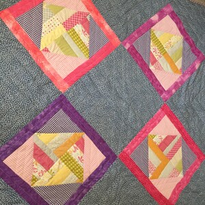 Puede incluir: Un edredón hecho a mano con cuatro bloques en forma de diamante. Cada bloque tiene un diseño central de patchwork en rosa, amarillo y verde, enmarcado por un patrón de cuadros y un borde rosa o morado liso. El fondo del edredón es un estampado floral azul.