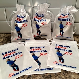 May include: Six white satin drawstring bags with a cowboy theme. Each bag has a printed image of a person wearing a cowboy hat and a blue shirt with the text "Cowboy Nikki Hangover Kit" and "44th Birthday".