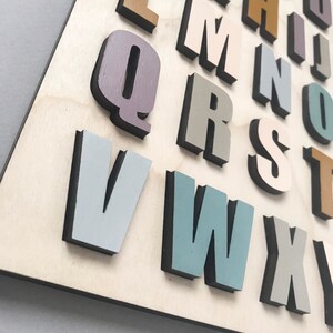 May include: A set of wooden alphabet letters painted in various pastel colors, including light blue, gray, beige, and brown. The letters are arranged in alphabetical order on a light wood background.