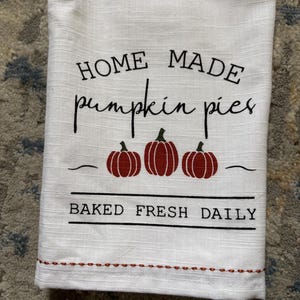 May include: White kitchen towel with the text "HOME MADE pumpkin pies" in black script. Three red pumpkins are below the text, and the words "BAKED FRESH DAILY" are printed below the pumpkins. A decorative brown stitched border is at the bottom.
