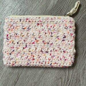 Könnte beinhalten: Eine weiße gehäkelte Reißverschlusstasche mit einem gesprenkelten Muster aus rosa, orange, gelben und violetten Garn.