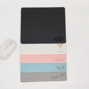 Könnte beinhalten: Ein Stapel von fünf personalisierten Mauspads mit verschiedenen Farben und Designs. Das oberste Mauspad ist schwarz mit einem goldenen Logo und dem Text "Michelle". Das zweite Mauspad ist weiß mit einem goldenen Tesla-Logo und dem Text "Tesla". Das dritte Mauspad ist rosa mit dem Text "Laura Moran". Das vierte Mauspad ist hellblau mit einem goldenen Kranz und dem Text "Happy". Das unterste Mauspad ist grau mit dem Text "Wendy Turner".