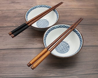 May include: Two wooden chopsticks and two white ceramic bowls with blue and white floral patterns. The bowls are on a brown wooden surface.