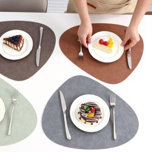 Puede incluir: Cuatro manteles individuales de forma irregular en marrón, gris y verde están dispuestos sobre una superficie blanca. Cada mantel tiene un plato blanco con un postre, un tenedor y un cuchillo. Una persona sostiene un plato con un postre.