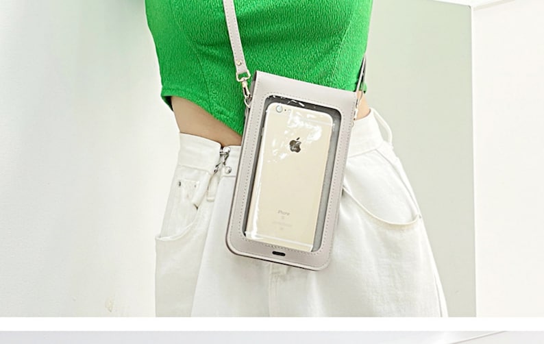 May include: A gray crossbody phone bag with a clear plastic window to view the phone screen. The bag has a silver chain strap and is worn over a white shirt.